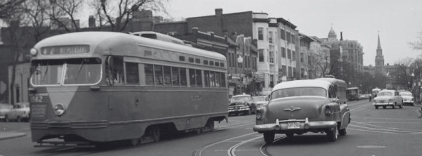 Black-and-white historic street scene with a streetcar, mid-century cars and rows of neighborhood buildings