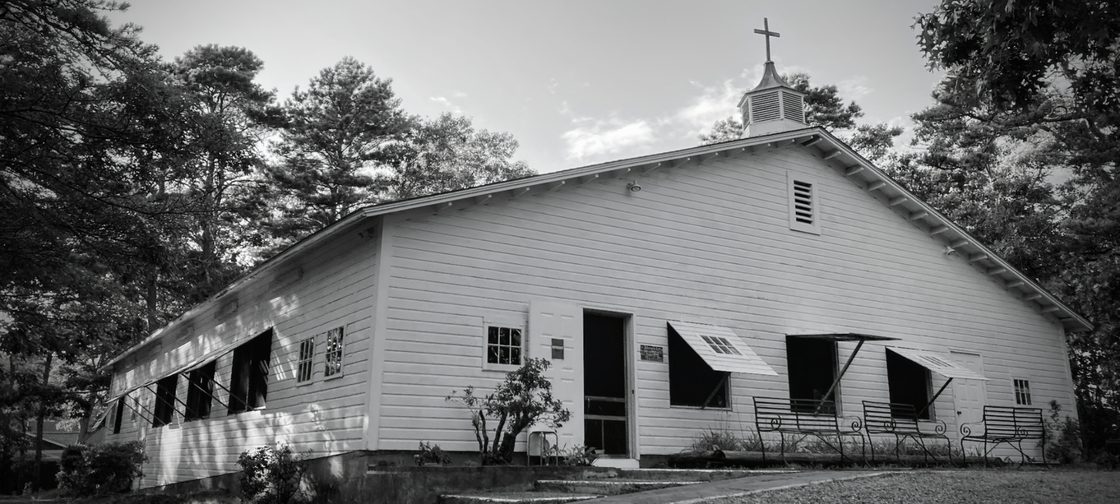 Black-and-white photograph of a white clapboard building at Onset Campground, with open windows, benches and a cross-topped cupola
